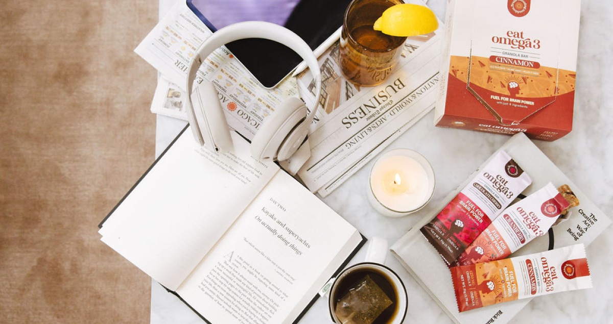 Flat lay image of a book, tea, headphones and newspaper and candle that showcases the Omega 3 granola bar trio.