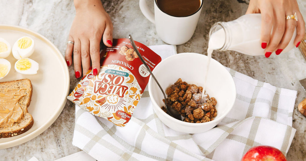 Flay lay image of a person's hands pouring milk into a bowl containing Omega 3 Mega O's Cinnamon Cereal Bites with other healthy food on the table.